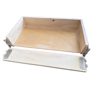 Puede incluir: Una caja de madera con una tapa con bisagras. La caja está abierta y la tapa está colocada plana. La caja está hecha de madera clara y tiene un acabado natural.
