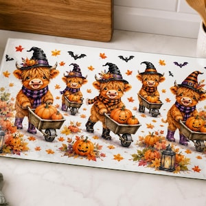 May include: A rectangular glass cutting board featuring a festive autumn illustration of five fluffy Highland cows wearing witch hats and scarves. The cows are pushing wooden wheelbarrows filled with orange pumpkins amidst falling maple leaves, bats, and glowing lanterns.
