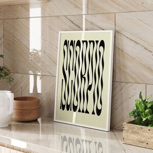 May include: A white framed print featuring a black and white graphic design of the word "Scorpio" on a light beige background. The print is displayed on a countertop alongside a white ceramic jug, a wooden bowl, and a wooden planter filled with green plants.