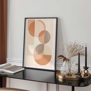 May include: An abstract art print in a black frame, displaying overlapping geometric shapes in beige, tan, and orange. The print is on a black table with candles, vases, and a tray. The style is modern and minimalist. The table is approximately 100 cm in length.