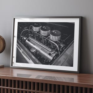 May include: Black and white photograph of a vintage car engine with three air filters. The engine is made of metal and has a lot of detail. The word "Ferrari" is visible on the engine.