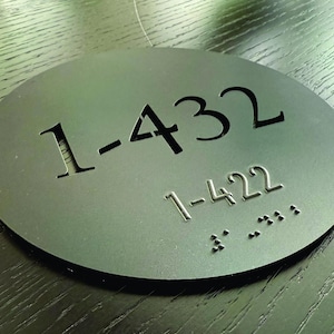 May include: A black, oval-shaped sign with the numbers "1-432" cut out. Below, the number "1-422" is printed in a smaller font, along with Braille dots. The sign is on a dark wood surface.