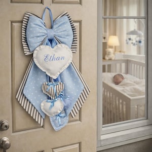 Può includere: Un fiocco per porta per bambini blu e bianco con un cuore centrale ricamato con il nome "Ethan". Il fiocco presenta un motivo a pois, dettagli a righe e ornamenti a forma di cuore sospesi con nastri blu. Un bambino dorme in una culla visibile attraverso una finestra.