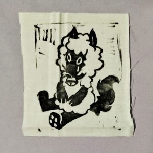 May include: A square fabric patch featuring a black ink block print of a stylized, fluffy creature with large eyes and a bushy tail. The design is centered on a plain, off-white woven textile square with visible fabric texture and frayed edges.