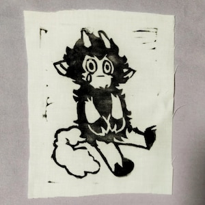 May include: A rectangular white fabric patch featuring a black ink screen-printed illustration of a small, furry creature with horns, large eyes, and a tear on its cheek. The creature is sitting down with a fluffy tail and dark-colored paws.