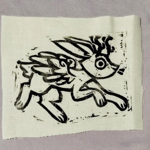 May include: A rectangular piece of off-white fabric featuring a black ink block print of a mythical creature resembling a rabbit with wings and antlers. The design is rendered in a bold, graphic style with visible texture from the printing process.