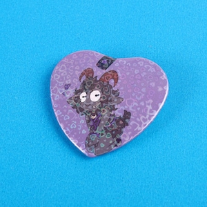 May include: Purple heart-shaped pin with a cartoon goat with horns and a purple and green patterned body. The background is a purple pattern of hearts.