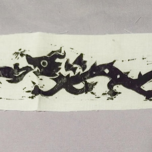 May include: A rectangular piece of off-white fabric featuring a black block-printed design of a long, serpentine creature with a large eye, jagged back, and a curled tail, oriented horizontally across the centre of the textile material.