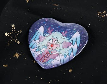 Könnte beinhalten: Herzförmiger Button mit einer skurrilen Illustration einer Kreatur mit Geweih, Flügeln und einem traurigen Gesichtsausdruck. Das Kunstwerk verwendet Blau-, Lila-, Rosa- und Weißtöne mit einem sternenübersäten Hintergrund. Der Button liegt auf schwarzem Stoff.