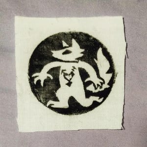 May include: A square fabric patch featuring a black circular design on an off-white background. The graphic depicts a stylized creature with pointed ears and a tail, centered within the circle, with a small heart shape on its chest area.