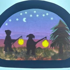 Può includere: Un'opera d'arte in carta ritagliata semicircolare che presenta la sagoma di un bambino e di un cane che camminano in un paesaggio notturno. Entrambe le figure portano bastoni con lanterne gialle luminose. La scena include luna e albero.