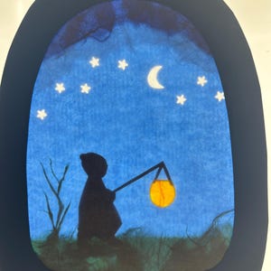 Può includere: Un'opera d'arte in carta ritagliata retroilluminata che mostra la sagoma scura di una figura incappucciata in piedi in un campo erboso di notte. La figura tiene una lanterna gialla luminosa. Il cielo blu presenta una luna crescente bianca.
