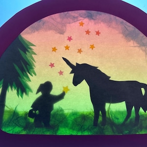 Puede incluir: Una obra de arte en papel recortado que muestra la silueta oscura de una persona y un unicornio en un campo con un árbol. El fondo es un degradado de verde, amarillo y rosa, decorado con pequeñas estrellas naranjas y rosas en el cielo.