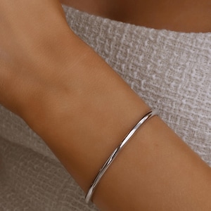Può includere: Un bracciale a cerchio minimalista in argento. La sottile fascia in metallo lucido si curva attorno al polso, riflettendo la luce. Il bracciale è un gioiello semplice ed elegante.