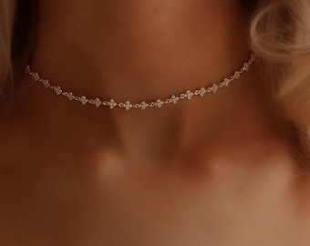 May include: A delicate gold-tone choker necklace featuring a continuous chain of small, four-petalled floral links. Each individual link is encrusted with tiny, clear, sparkling stones that catch the light, creating a subtle, shimmering effect around the neck.