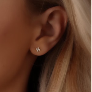 May include: A close-up of a small, flower-shaped stud earring with multiple small, clear stones. The earring is set in a gold-colored metal and is worn on a person's ear. The person has blonde hair.