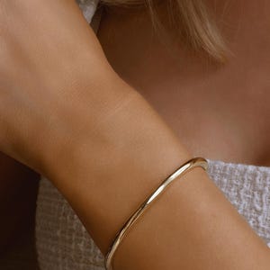 以下が含まれることがあります： ゴールドカラーのバングルブレスレット。ブレスレットは、丸みを帯びた形状のシンプルで滑らかなバンドです。ブレスレットは腕に着用されており、肌の色は明るいです。背景はぼやけています。