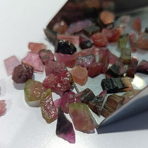 以下が含まれることがあります： ピンク、グリーン、ブラックのトルマリンの宝石のコレクション。宝石はさまざまな形とサイズで、白い表面に散らばっています。