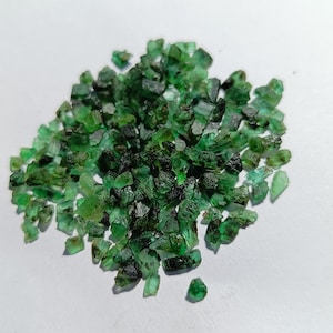May include: A pile of raw, uncut emerald gemstones. The emeralds are a deep, rich green color, with varying shades and textures. The gemstones are scattered on a white surface, showcasing their natural, rough appearance. These gemstones are ideal for jewelry making.