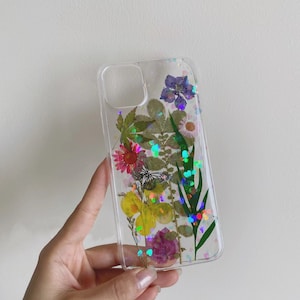 May include: A clear phone case with pressed flowers and iridescent glitter. The flowers include pink, yellow, purple, and green.