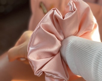 May include: A soft, pink satin scrunchie for hair. The scrunchie is made of a smooth, silky fabric and is designed to be gentle on hair.