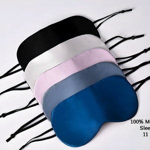 May include: Six silk sleep masks in different colours, including black, white, pink, blue, and grey. The masks are arranged in a stack with the straps hanging down. The text "100% Mulberry Silk Sleep Mask 11 Colours" is visible at the bottom of the image.