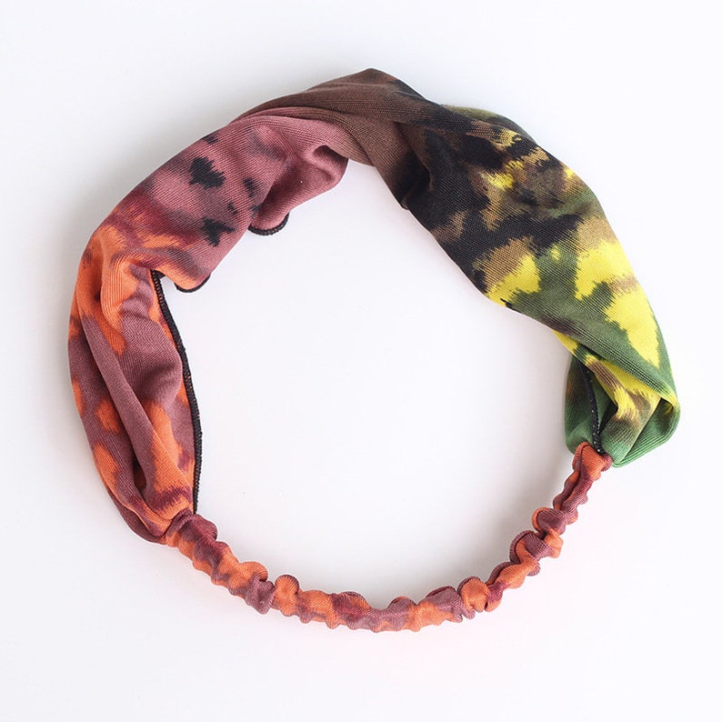 Może przedstawiać: Elastyczna opaska do włos&oacute;w z węzłem. Opaska wykonana jest z miękkiej tkaniny z wzorem tie-dye w odcieniach pomarańczowego, brązowego, zielonego i ż&oacute;łtego.