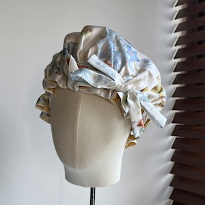 May include: A white satin shower cap with a floral print in shades of blue, yellow, and orange. The cap has a gathered ruffle around the edge and a bow on top.