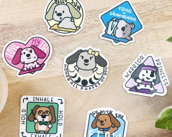 May include: A collection of colorful dog-themed stickers with various designs and text. The stickers feature cartoon dogs in different poses, with phrases like "Hello Sun," "Vibing Shavasana," and "Warrior Princess." The stickers are on a wooden surface.