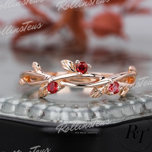 May include: A rose gold ring with a nature-inspired design. The ring features a delicate vine motif with small leaves and three vibrant red gemstones. The ring is displayed on a textured surface with the text "RollinsTeus Art Deco Jewelry".
