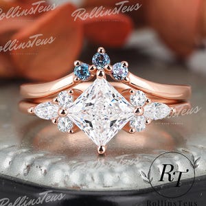 Puede incluir: Un anillo de compromiso de oro rosa con un diamante cuadrado de talla princesa y una banda delicada adornada con diamantes más pequeños y piedras preciosas azules. El anillo está colocado sobre una superficie texturizada.
