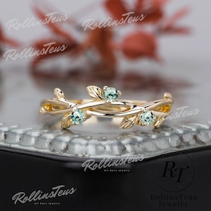 May include: A delicate gold ring with a vine design and three small green gemstones. The ring is on a dark grey surface.