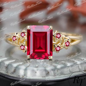May include: A gold ring with a large, rectangular, red gemstone at its centre. Small red gemstones are set in star-shaped designs on either side of the main stone. The ring has a delicate, openwork design.