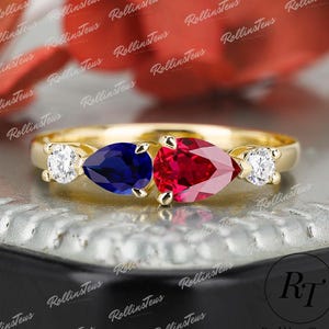 Può includere: Un anello in oro con uno zaffiro blu centrale taglio a pera e un rubino rosso taglio a pera, affiancati da due diamanti bianchi taglio rotondo. La fascia è in oro lucido e le pietre sono disposte orizzontalmente sulla parte superiore.