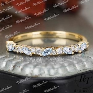 Puede incluir: Un anillo de banda de oro con piedras preciosas alternas de color azul claro en forma de marquesa y redondas blancas. El anillo está sobre una superficie oscura. El texto "RollinsTeus Jewelry" es visible.