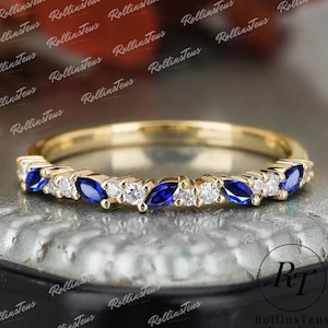 Puede incluir: Un anillo de banda dorado con piedras preciosas azules en forma de marquesa y piedras blancas redondas alternas. El anillo está sobre una superficie reflectante. Es una joya.