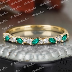 Puede incluir: Un anillo de banda de oro con esmeraldas en forma de marquesa y diamantes blancos redondos alternados. El anillo tiene un diseño delicado, con las piedras preciosas muy juntas. El anillo se muestra sobre una superficie texturizada.