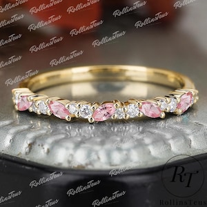 Puede incluir: Un anillo de banda de oro con piedras preciosas rosas y blancas alternas. El anillo presenta piedras rosas con corte marquesa y piedras blancas redondas. El anillo se muestra sobre una superficie reflectante.