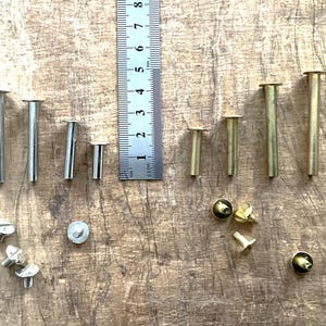 Könnte beinhalten: Eine Auswahl an Metallnieten und Schrauben in Silber- und Messingtönen, angeordnet auf einer Holzoberfläche. Die Nieten variieren in der Länge, die längste misst etwa 7,6 cm. Ein Lineal liegt zur Größenbestimmung daneben.
