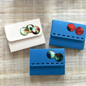 Könnte beinhalten: Drei kleine, handgefertigte Papiergeldbörsen in Weiß und Blau. Jede Geldbörse hat ein einzigartiges Design aus farbenfrohen, kreisförmigen Verzierungen. Die Geldbörsen sind auf einer hellen Holzoberfläche angeordnet.