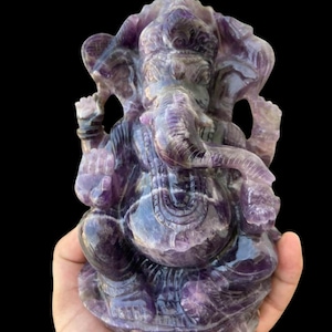 May include: Purple amethyst Ganesha statue carved in detail. The statue is sitting on a base and has a trunk raised in the traditional blessing pose.