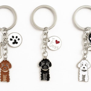 May include: Three dog-themed keyrings with silver rings. Each keyring features a dog charm, a bone charm, and a circular charm with a paw print or the text "I Love Dogs". The dog charms are brown, black, and white.