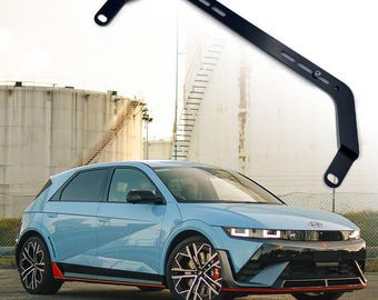 May include: A black metal car strut bar is shown above a light blue Hyundai Ioniq 5. The car has black and silver wheels with red detailing. The strut bar has mounting holes and is designed to improve vehicle handling.