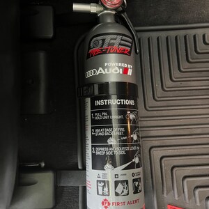 May include: A silver fire extinguisher with a black handle and a gauge on top. The extinguisher has a black label with the text "The Tuner" and "Powered by Audi". The label also includes instructions for using the extinguisher.