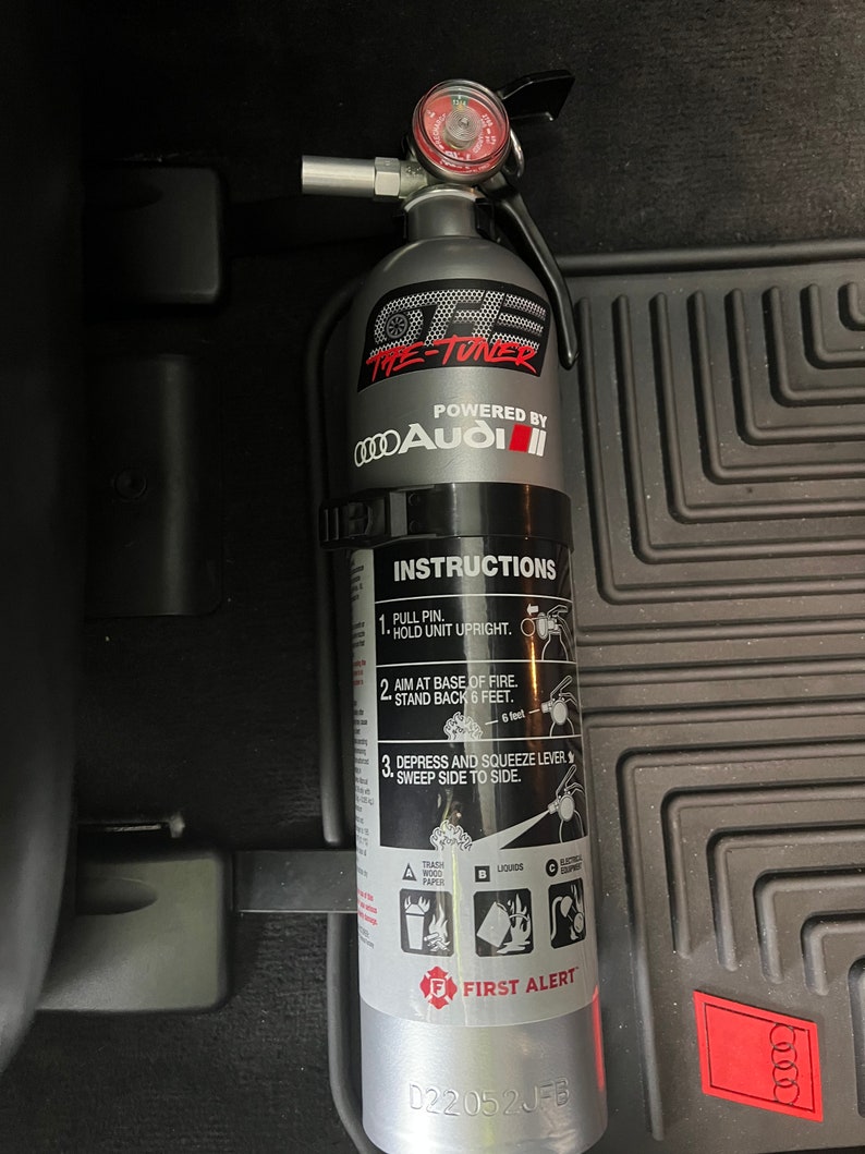 May include: A silver fire extinguisher with a black handle and a gauge on top. The extinguisher has a black label with the text "The Tuner" and "Powered by Audi". The label also includes instructions for using the extinguisher.