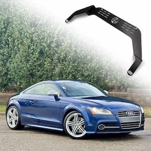 Puede incluir: Un cupé Audi TT azul con llantas plateadas está estacionado en una carretera. Encima del coche, una abrazadera de metal negro con un logotipo y ranuras. El fondo presenta un seto verde y un cielo borroso.