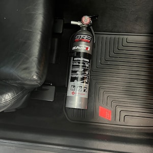 May include: A silver fire extinguisher with a red gauge and a black handle. The extinguisher is labeled "GTE Fire-Toner" and "Powered by Audi". The extinguisher is mounted in a car.