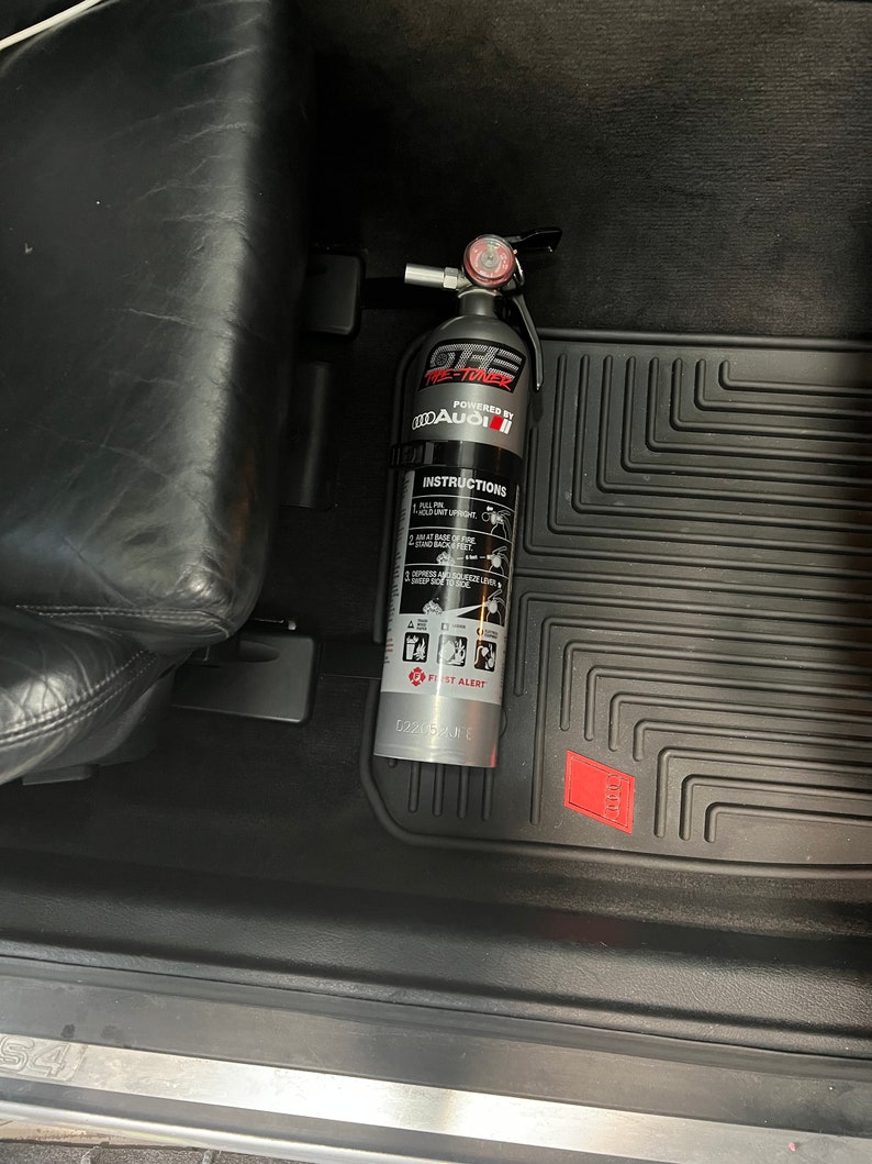 May include: A silver fire extinguisher with a red gauge and a black handle. The extinguisher is labeled "GTE Fire-Toner" and "Powered by Audi". The extinguisher is mounted in a car.
