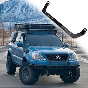 Op de afbeelding: Een blauwe SUV met een dakdrager, terreinbanden en een zwarte snorkel. Een zwarte metalen beugel is rechtsboven afgebeeld. Het voertuig staat geparkeerd voor een berg.