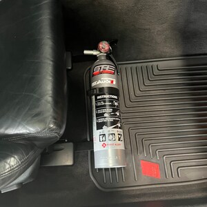 May include: A silver fire extinguisher with a black handle and a red gauge. The extinguisher is labeled "OFF-ROAD", "FIRE-TANK", "POWERED BY", "ODDAUGI", and "FIRST ALERT". The extinguisher is mounted in a car.